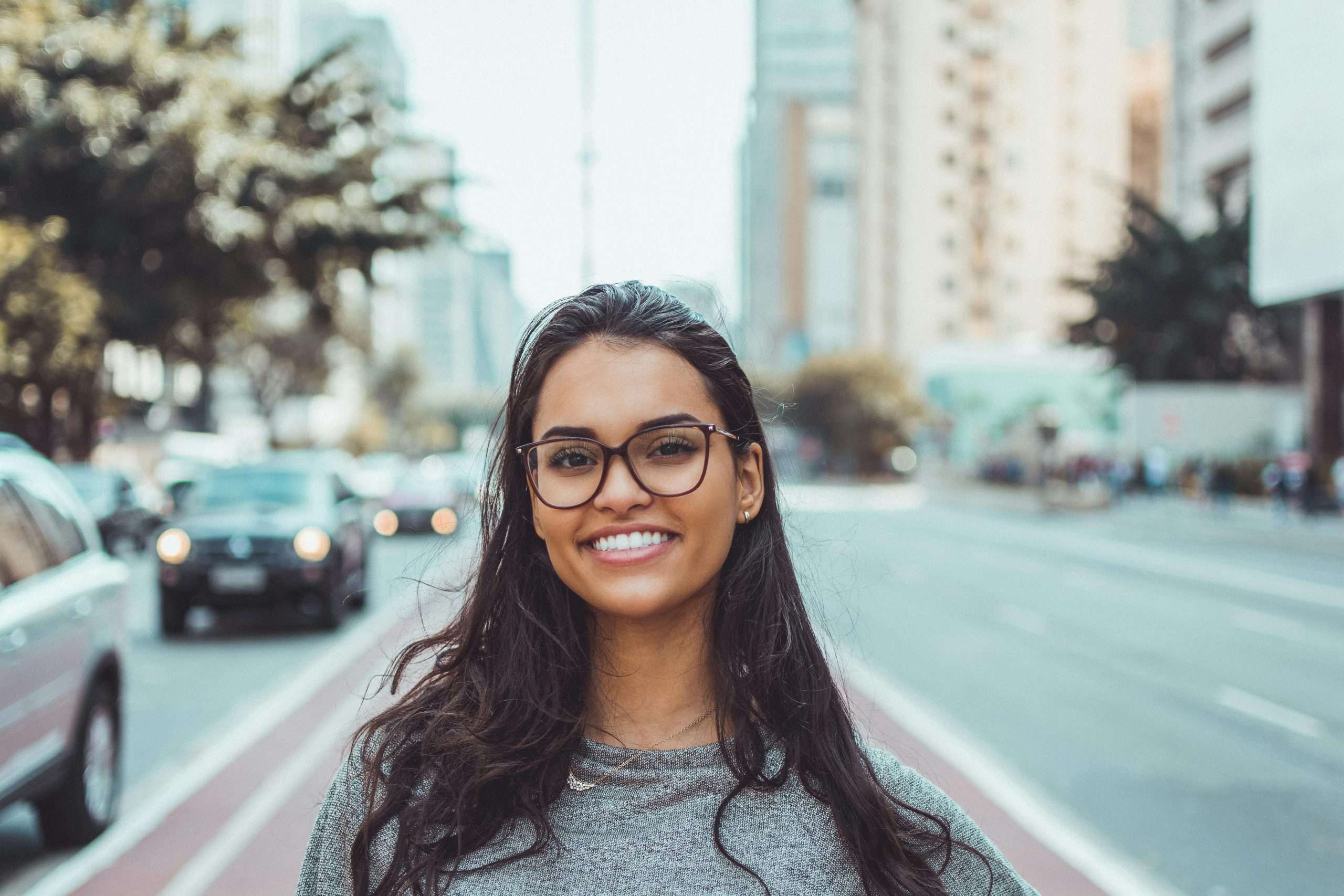 pexels-photo-1239291-1239291 A young woman with glasses smiling on a city street, embracing urban lifestyle.